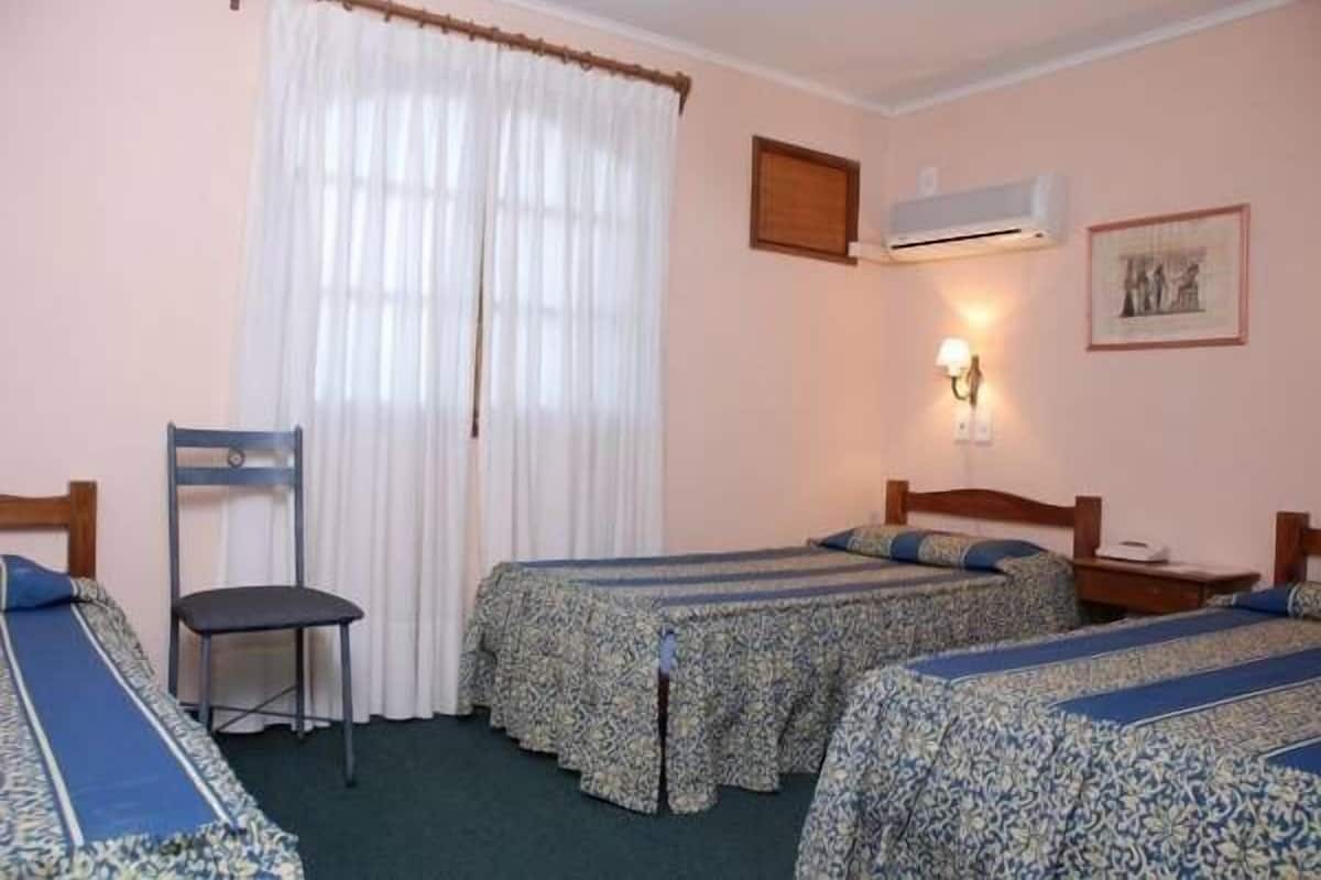 A room with two single beds, a chair, a window with curtains, an air conditioning unit, and a wall-mounted lamp.