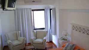 Une chambre d’hôtel avec un lit, deux fauteuils, une petite table avec une lampe et une fenêtre avec des rideaux.