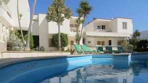 Una piscina con sillas de descanso, rodeada de edificios blancos con balcones y palmeras.
