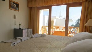 Habitación de hotel con cama, mesita de noche con una botella y un vaso, teléfono y vistas al balcón exterior.