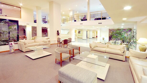 A spacious, well-lit hotel lobby with high ceilings, multiple seating areas, and a large indoor plant.