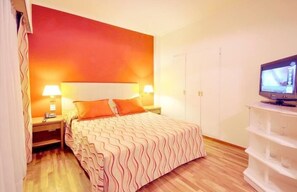Una habitación de hotel con cama, dos lámparas de noche, televisor y una estantería blanca.