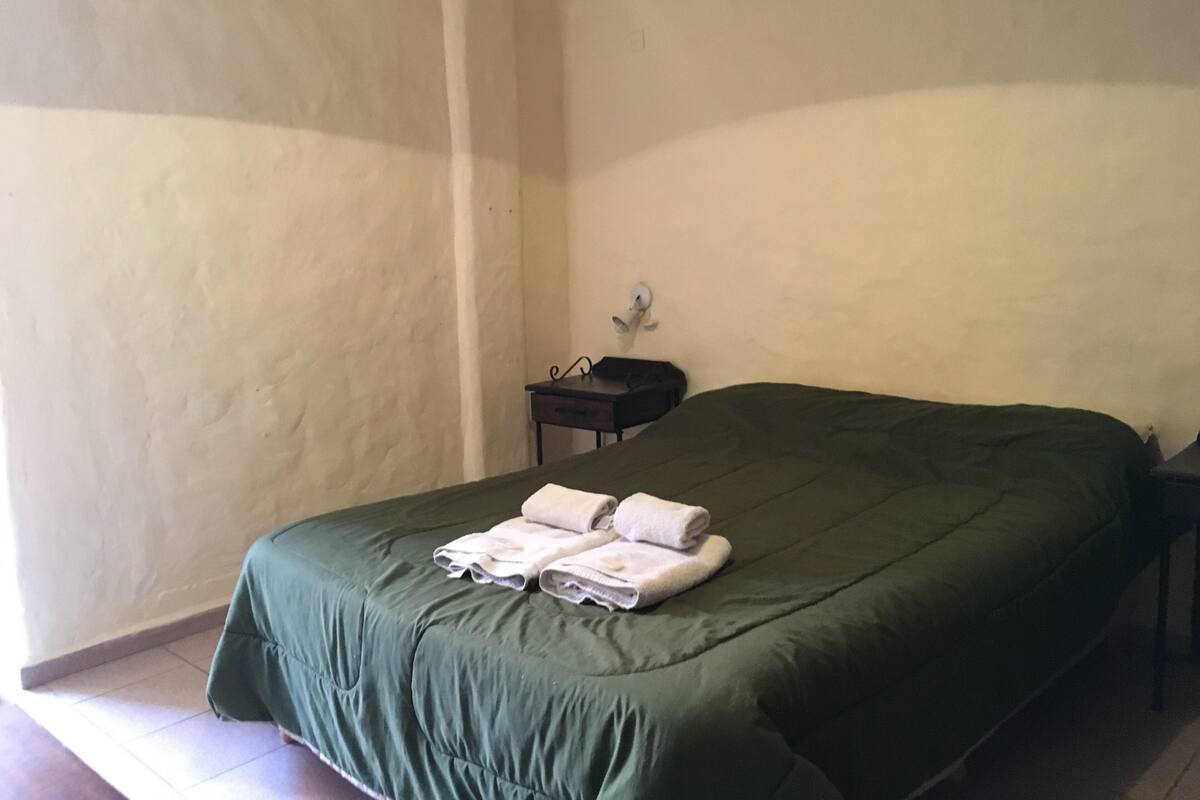 A single bed with a green bedspread and white towels folded on it. There is a small bedside table with a lamp and a wall-mounted light fixture.