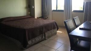 Quarto de hotel com uma cama, uma mesa com cadeira, uma luminária e uma janela com cortinas.