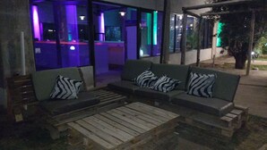 Uma área externa com assentos, incluindo um sofá e mesas de madeira, um banco e uma porta de vidro que dá para um espaço interno com iluminação roxa.