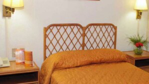 A hotel room with a wooden headboard, a bed with an orange bedspread, a bedside table with a lamp, a vase with flowers, and a curtain on the window. - Hotel Miranda Cumberland  (Santa Ana)