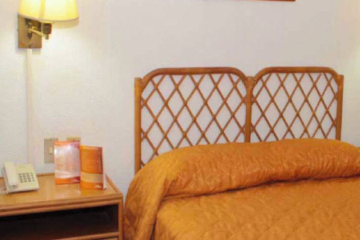 A hotel room with a wooden headboard, a bed with an orange bedspread, a bedside table with a lamp, a vase with flowers, and a curtain on the window.