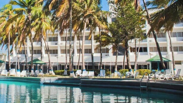 A swimming pool with lounge chairs and umbrellas, surrounded by palm trees and a hotel building. - Hotel Miranda Cumberland  (Santa Ana)