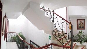 Un escalier avec des rampes rouges et un motif décoratif en métal. On y voit une porte avec un numéro, un tableau encadré accroché au mur et une plante en pot sur le palier.
