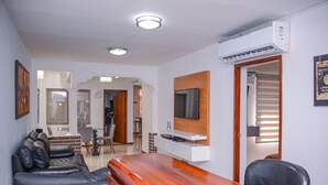 A modern living room with a wooden bar, a leather sofa, a TV mounted on the wall, and a dining area in the background. - Brown Stone Apartments (Medellín)