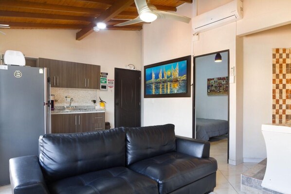 A modern living room with a black leather sofa, a kitchen with dark cabinets, and a painting of a sailboat on the wall.