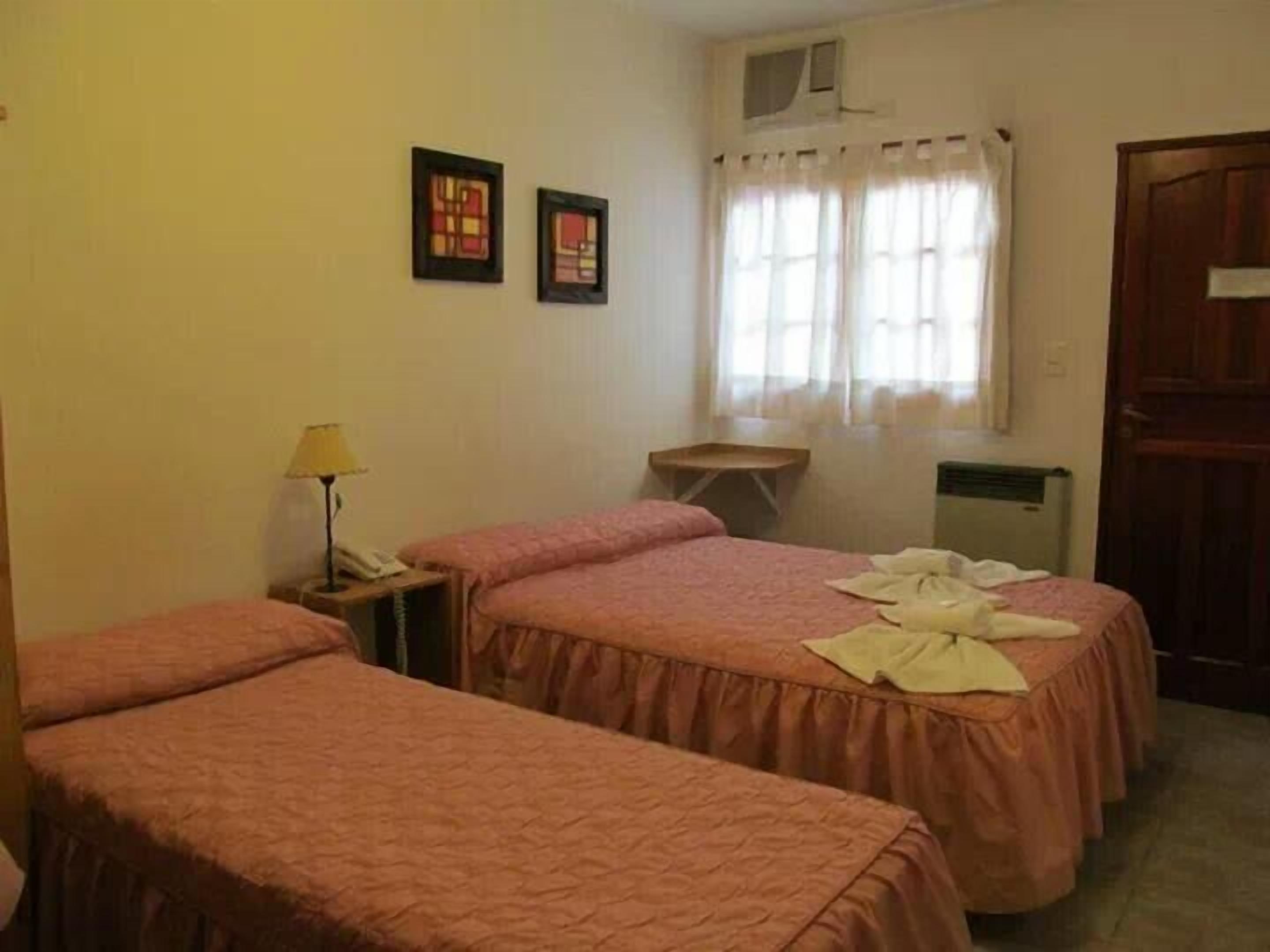 Quarto de hotel com duas camas, uma mesinha com abajur, uma janela com cortinas e uma porta.