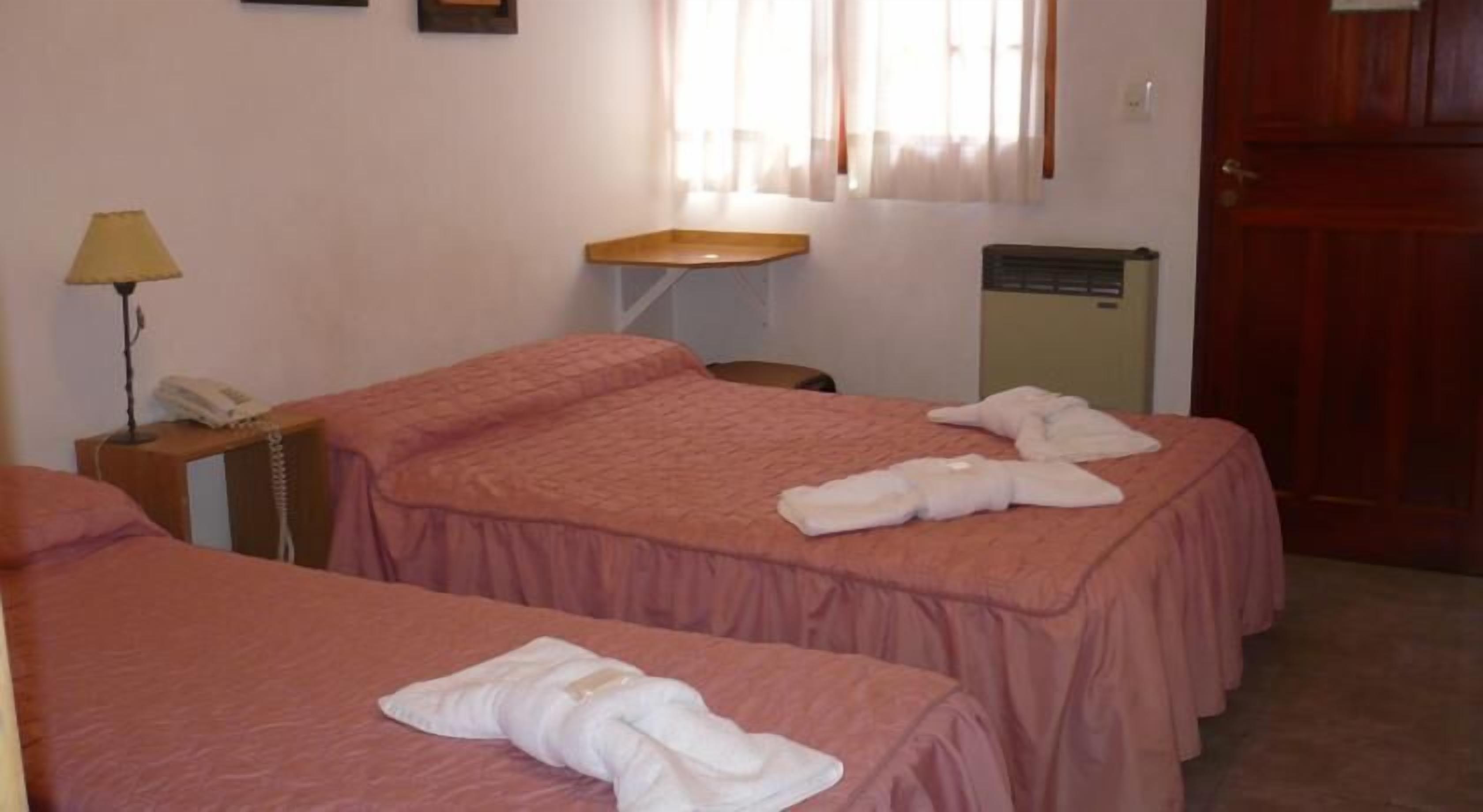 Una habitación de hotel con dos camas, una mesita de noche con l ámpara, una mesita pequeña y una ventana con cortinas.