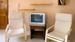 Una habitación con dos sillones blancos acolchados, una pequeña mesa de madera con un televisor, y una estantería de madera encima de la mesa.