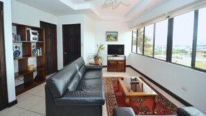 Ein Wohnzimmer mit einem schwarzen Ledersofa, einer Holzunterhaltungsecke, einem Fernseher und einem großen Fenster mit Blick auf die Stadt.
