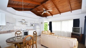 Eine moderne Küche mit weißen Schränken, einem Esstisch mit Stühlen, einem beigen Sofa, einem Fernseher und einem Deckenventilator.