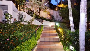 Eine Treppe, umgeben von  üppigem Grün und blühenden Pflanzen, die zu einem Gebäude mit weißer Fassade und Holzgeländer führt.