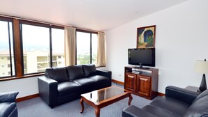 Una sala de estar moderna con un sofá de cuero negro, una mesa de centro de vidrio y un televisor de pantalla plana sobre un mueble de madera. Amplios ventanales ofrecen vistas a la ciudad.