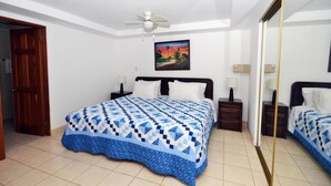 Un dormitorio con una cama, dos mesitas de noche, una mesita auxiliar, un ventilador de techo, un cuadro y una puerta.