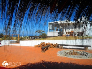 A patio area with a fire pit, a stack of firewood, and a building in the background.