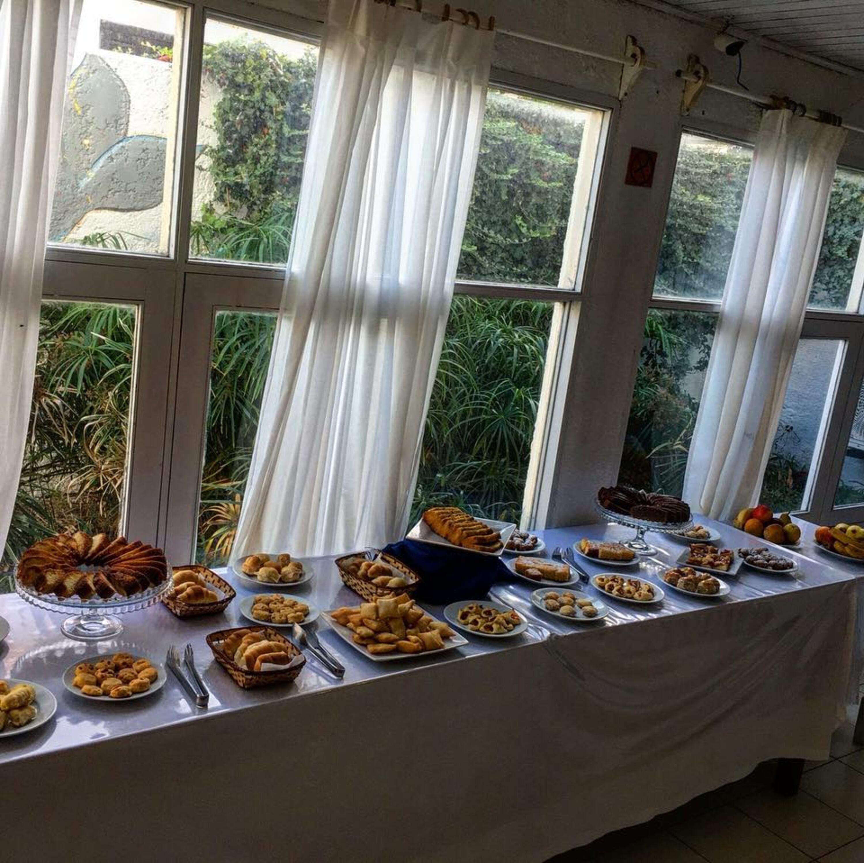 Una mesa de buffet con diversos alimentos, con un fondo de ventanas grandes con cortinas que dan a un jardín.