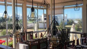 Un espace de restauration en plein air avec des tables dressées pour le repas, de grandes fenêtres offrant une vue sur le paysage environnant et des lampes suspendues.