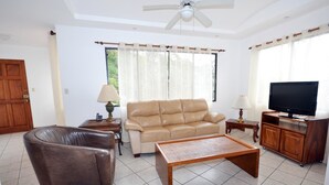 Un salon avec un canapé beige, un fauteuil marron, une table basse en bois, un téléviseur posé sur un meuble en bois et un ventilateur de plafond.