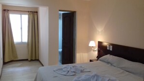 A hotel room with a bed, a nightstand with a lamp, a window with curtains, and an open door leading to another room.
