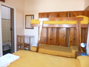 Uma cama beliche com cabeceira de madeira e roupa de cama amarela. Há uma pequena mesa de madeira e uma cadeira ao lado da cama. Um espelho está fixado na parede acima da cama.