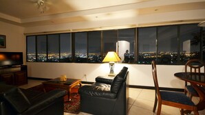 A modern living room with a city view, a flat-screen TV, leather sofa, and a dining area with wooden chairs. - Casa Reflejos Apartamento 9 (Escazu)