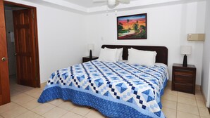 Un dormitorio con cama, dos mesitas de noche, ventilador de techo, una puerta y un cuadro colgado en la pared.