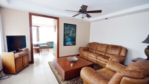 Una sala de estar con un sofá de cuero, una mesa de centro, un televisor sobre un mueble de madera y un ventilador de techo.