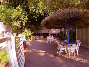 An outdoor seating area with tables and chairs, surrounded by lush greenery and a thatched umbrella.