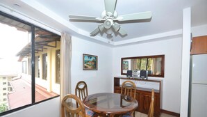 Un comedor con una mesa de vidrio redonda, sillas de madera, un ventilador de techo, una pequeña cocina con refrigerador y un ventanal con vistas a edificios.