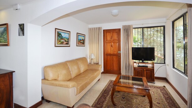Una sala de estar con un sofá color crema, una mesa de centro de madera, un televisor sobre un mueble bajo y un ventanal con cortinas.