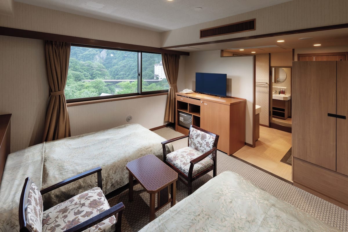 Ein Hotelzimmer mit zwei Betten, einem Schreibtisch mit Fernseher, einem Sessel und einem durchgegangenes Badezimmer.