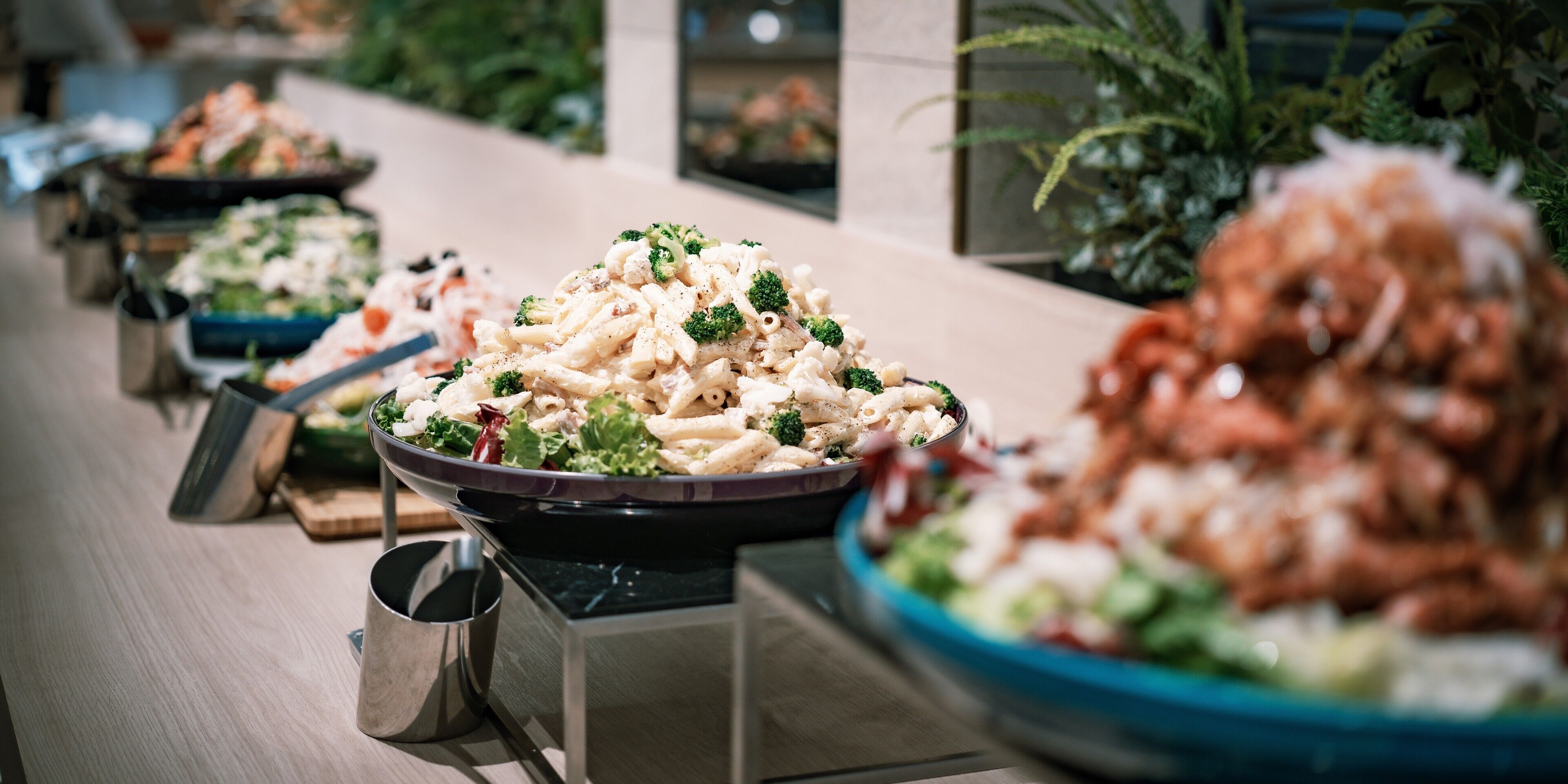 Ein Buffet mit verschiedenen Speisen, darunter Pasta und Salate, präsentiert auf einem langen Tisch mit Metall-Servierbesteck.