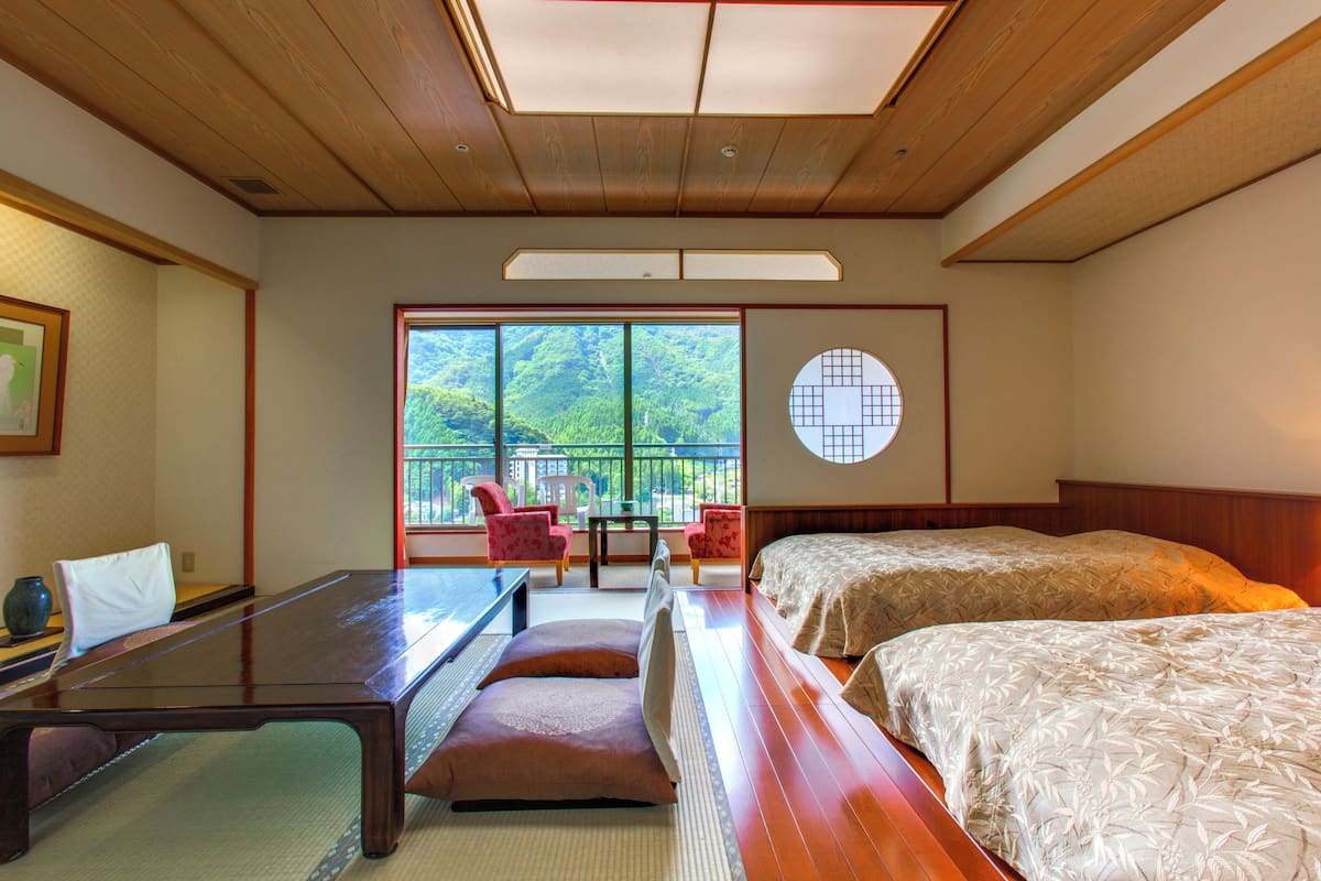 Een traditionele Japanse kamer met tatami vloeren, een lage houten tafel, twee bedden met tatami matten, en een groot raam met uitzicht op groen.