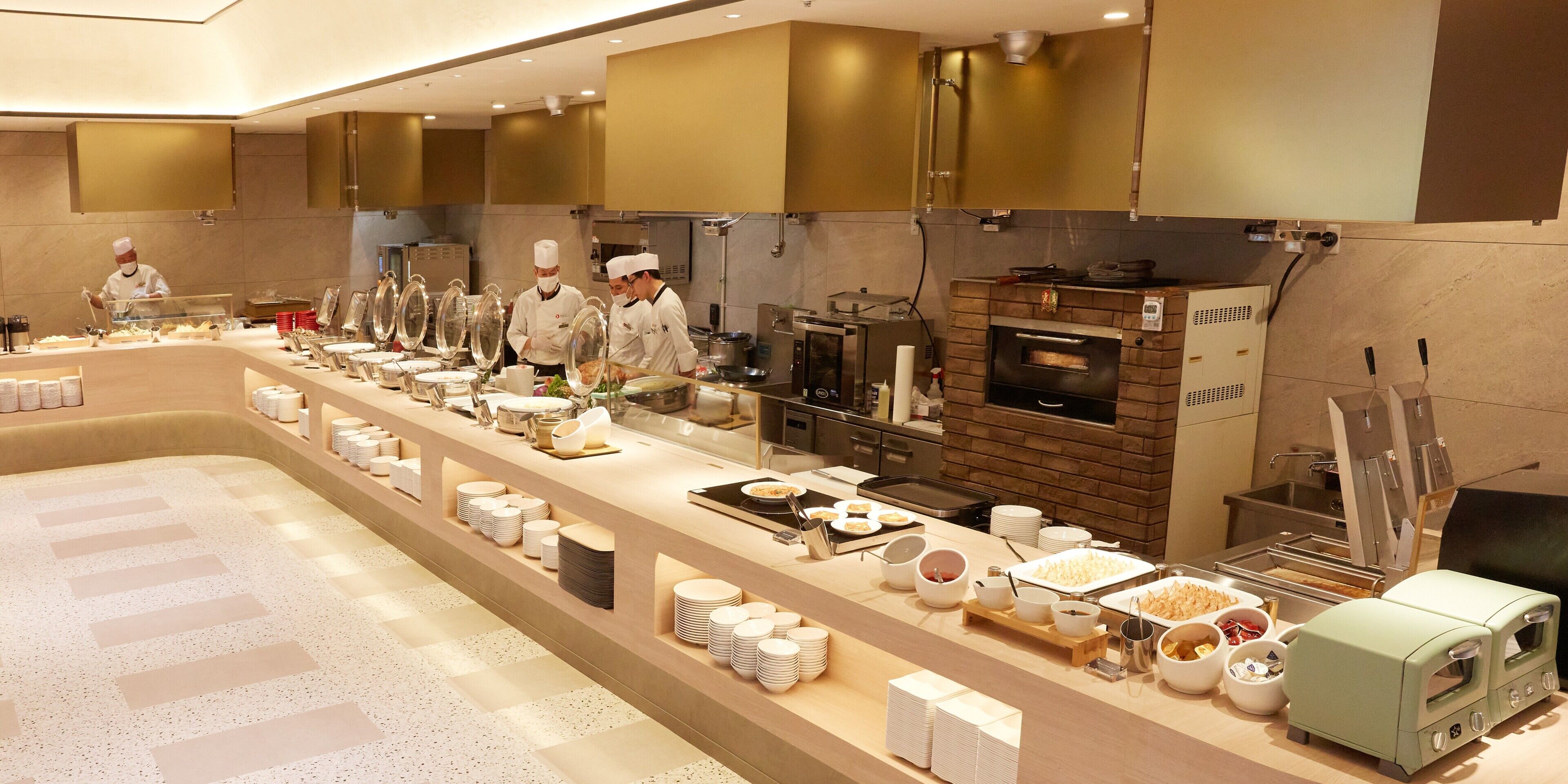 A modern kitchen with chefs preparing food, a brick oven, and a buffet line with various dishes.