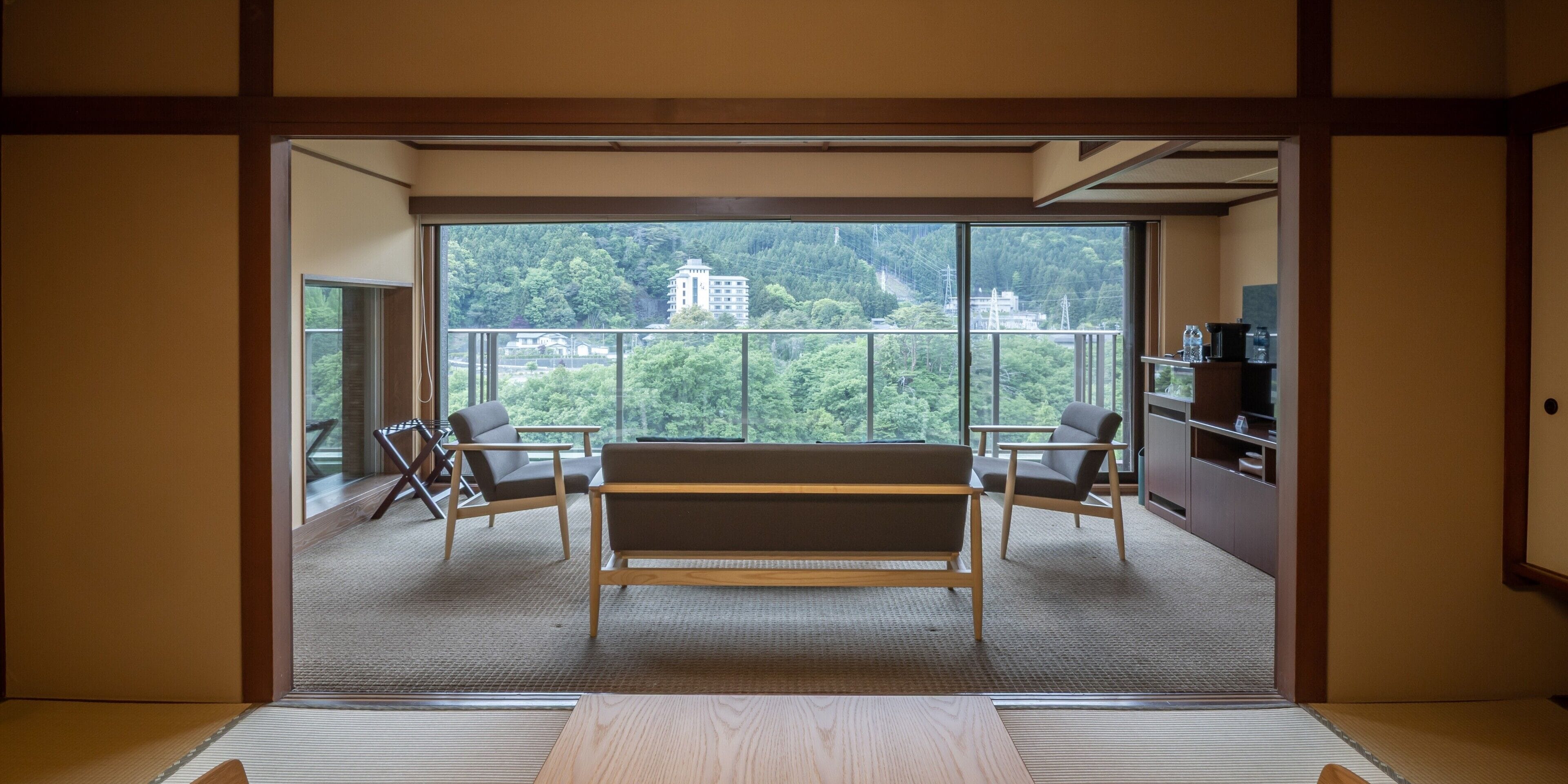 Ein Zimmer mit Tatami-Böden, einem niedrigen Holztisch, zwei Stühlen, einem Sofa und einem großen Fenster mit Blick auf ein Gebäude und Bäume.