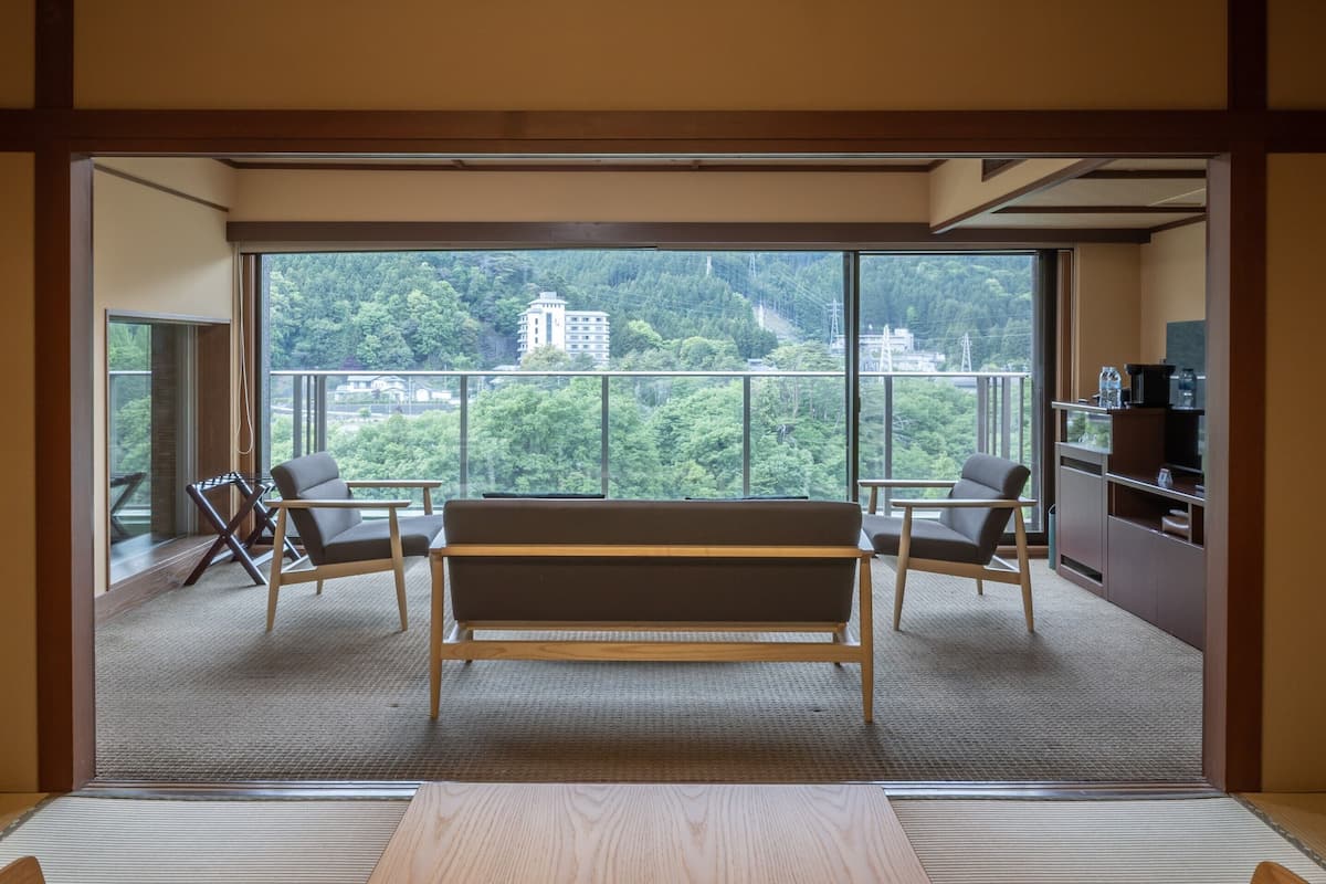 Ein Zimmer mit Tatami-Böden, einem niedrigen Holztisch, zwei Stühlen, einem Sofa und einem großen Fenster mit Blick auf ein Gebäude und Bäume.