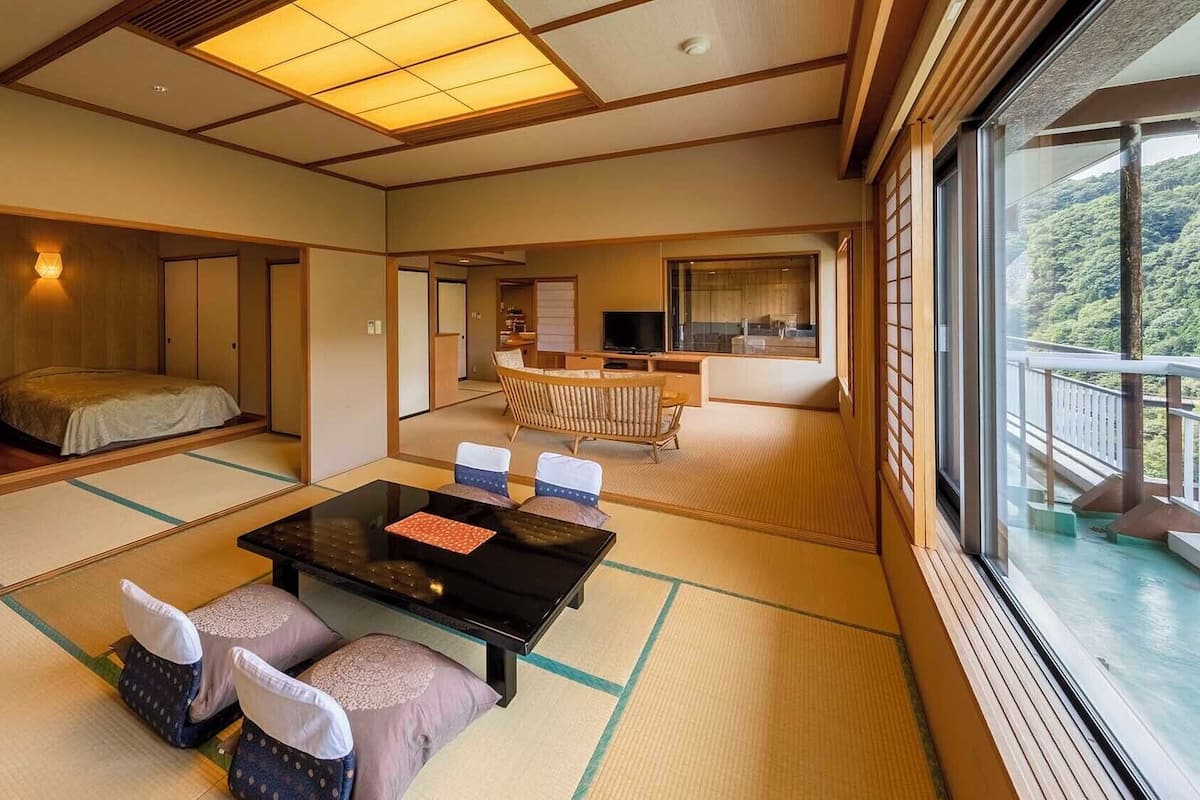 Een traditionele Japanse kamer met tatami vloeren, een salontafel met kussens, een bank en uitzicht op een weelderig bos.