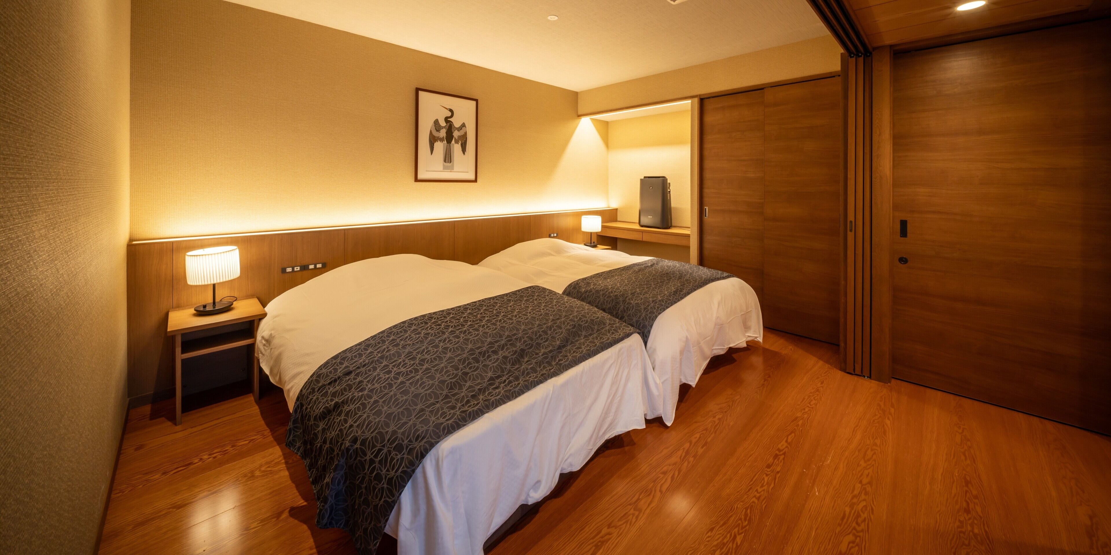 A hotel room with two beds, a nightstand with a lamp, a wooden floor, a framed picture on the wall, and a sliding door.