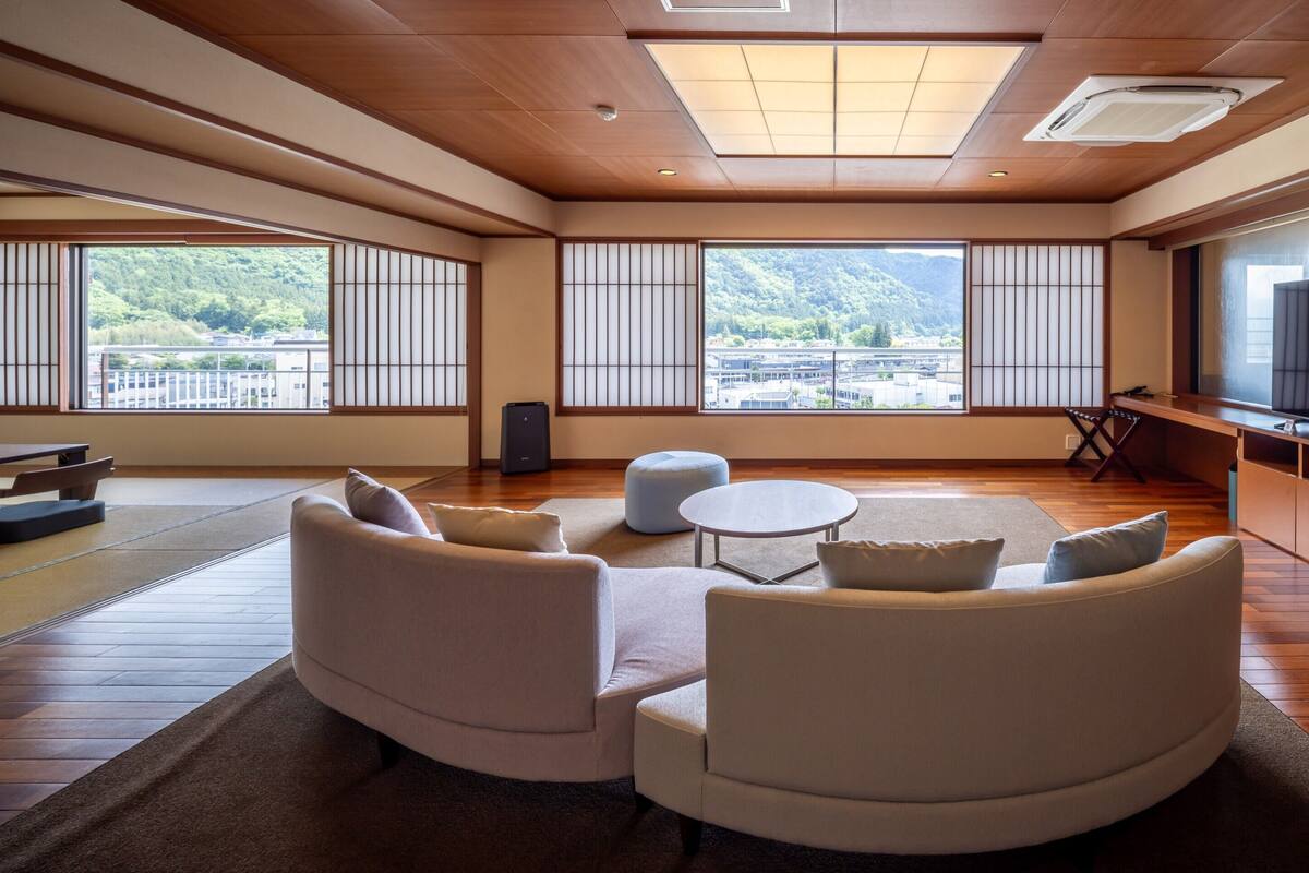 Ein modernes Wohnzimmer mit einer Sofagarnitur, einem Couchtisch, einem Fernseher und großen Fenstern mit Blick auf ein bewaldetes Gebiet.