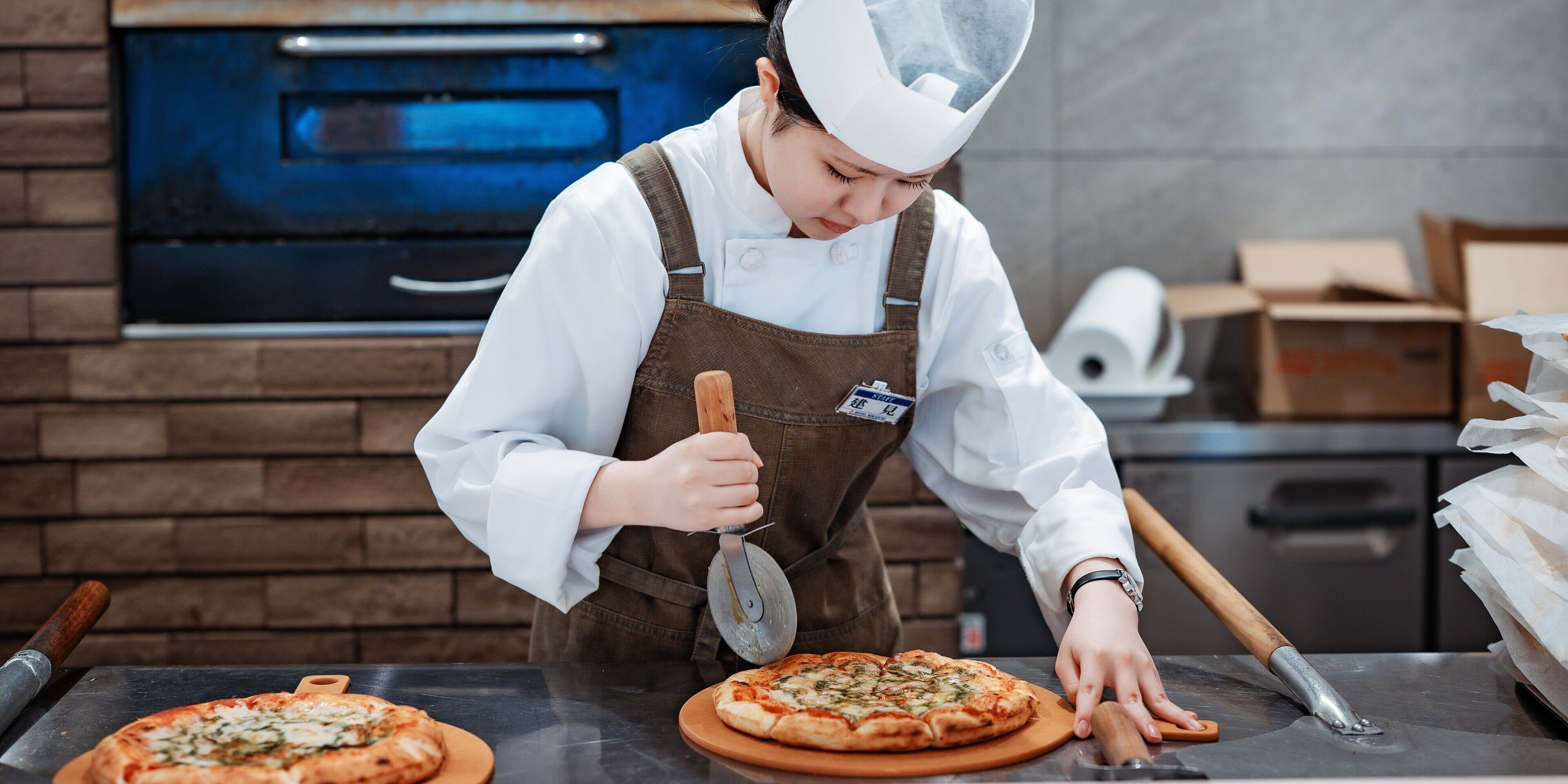 A person in a chef's uniform preparing pizza in a kitchen with a brick wall and a blue oven in the background.