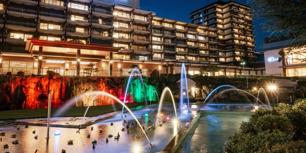 Une vue nocturne d’un hôtel à plusieurs étages, avec une fontaine et un espace piscine.