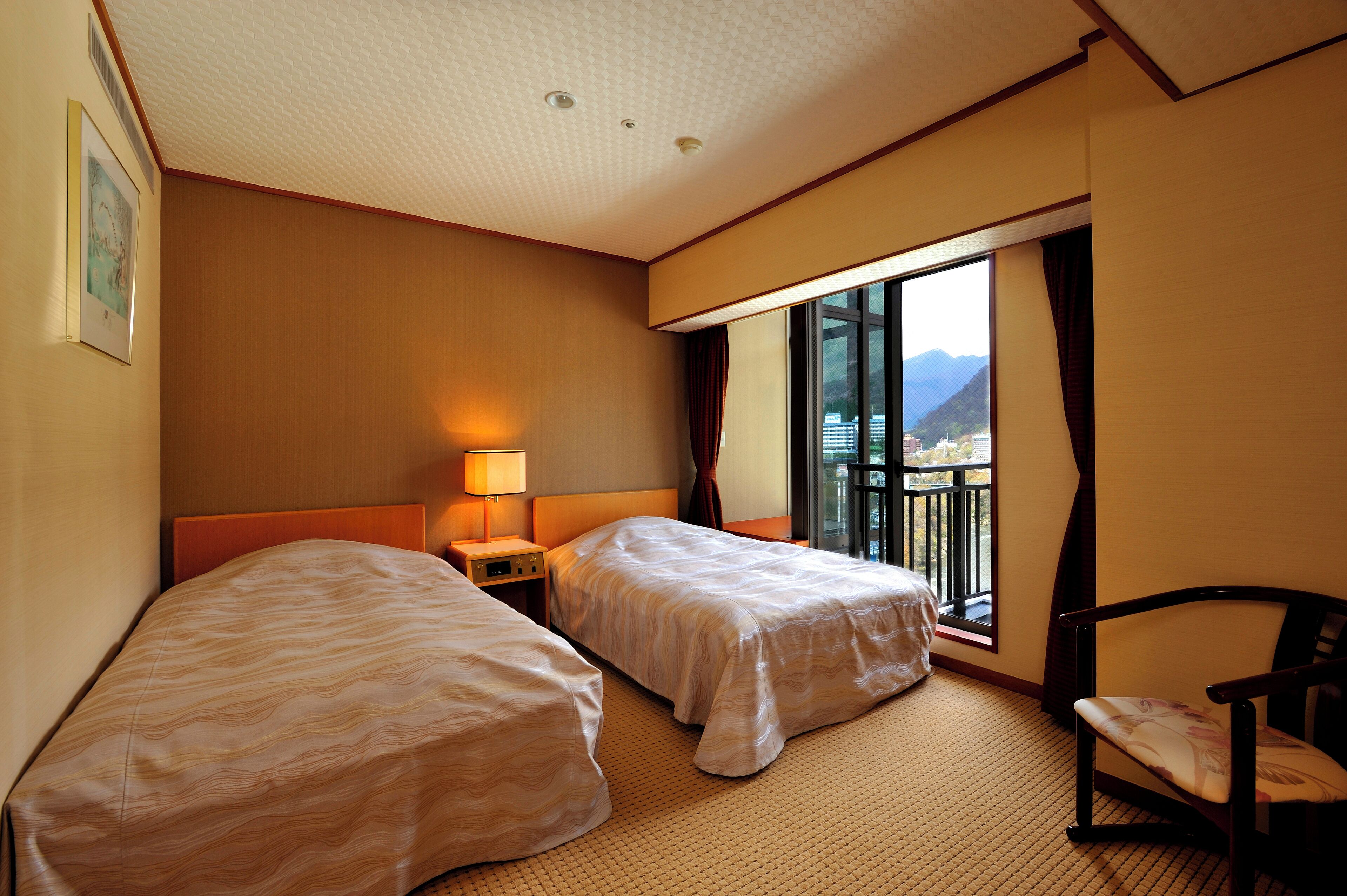 Habitación de hotel con dos camas, una mesita de noche con lámpara, una silla y un amplio ventanal con vistas a las montañas.