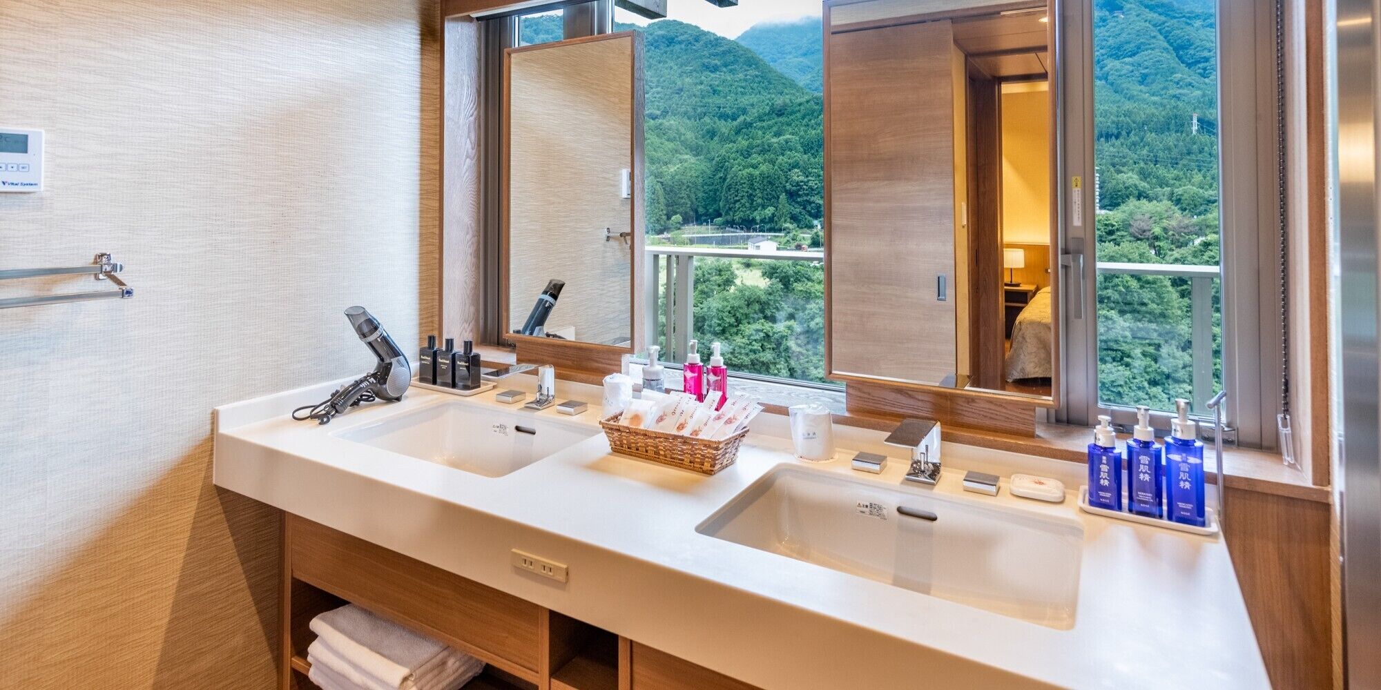 A bathroom with two sinks, a mirror, and a window with a view of a forested area.