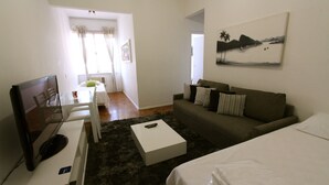 Una sala de estar moderna con televisor de pantalla plana, una mesa de centro blanca, un sofá gris y una cama con edredón blanco.