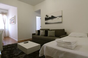 Habitación de hotel con cama, sofá, mesa de centro, un cuadro en la pared y ventana con cortinas.